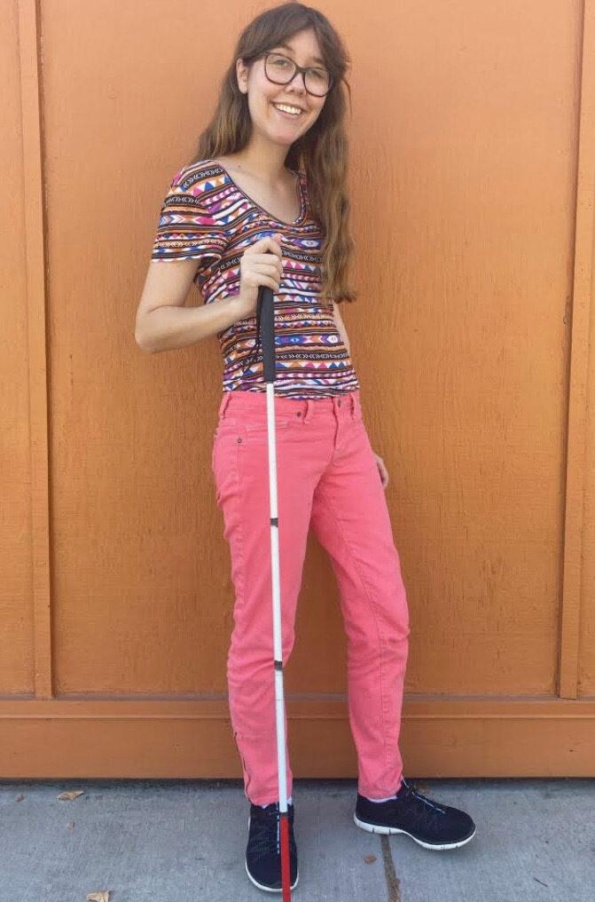 a young woman wearing glasses, a multicolored shirt, and pink pants poses with a cane against a wooden wall